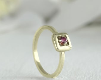 May include: A gold ring with a square setting featuring a small pink gemstone.