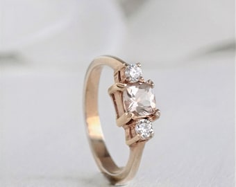 May include: A rose gold engagement ring with a square-shaped center stone and two smaller round stones on either side.