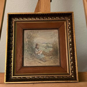 May include: A framed watercolor painting depicting a woman sitting in a field, sewing. The painting is set in a rural landscape with trees and a distant view of a village. The frame is gold with a brown inner border.