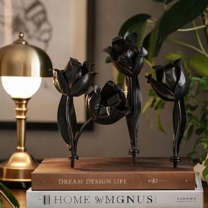 May include: Black metal tulip sculptures arranged on a stack of books with the titles "DREAM DESIGN LIFE" and "HOME & MGNUS". A gold-colored lamp with a white globe is visible in the background.
