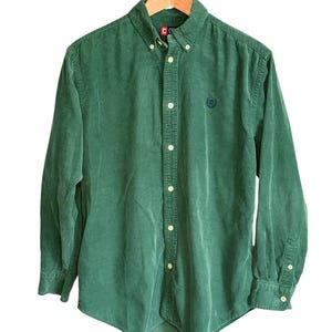 May include: A green corduroy button-down shirt with a button-down collar and long sleeves. The shirt has a small embroidered logo on the chest and is hanging on a wooden hanger.
