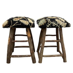 May include: Two stools with four legs made of rough, natural brown tree branches. Each stool features a square seat with a thick, textured hooked rug top in black, cream, and muted green floral patterns, supported by horizontal branch rungs.