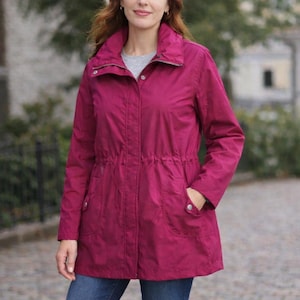 May include: A magenta-colored jacket with a stand-up collar, zipper closure, and drawstring waist. The jacket has two front pockets and long sleeves. The model is wearing a gray shirt underneath and blue jeans.