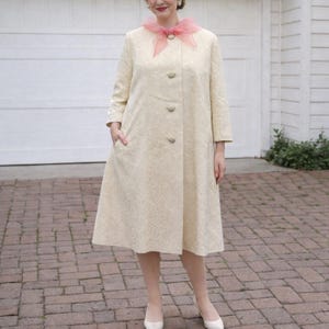May include: A cream-coloured vintage coat with a floral pattern, three buttons, and pockets. The coat is paired with white heels and a pink scarf. The coat has a knee-length hemline and three-quarter sleeves.