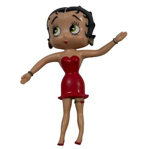 May include: A figurine of a cartoon character in a red dress with a heart-shaped bodice and a ruffled hem. The figure has black hair, green eyes, and is posed with one arm raised. The figurine has a garter with a red heart.