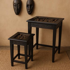 May include: Two black wooden tables with carved tops and decorative aprons. The tables are of different heights, with the taller one having a rectangular top and the shorter one a square top. Two wooden masks hang on the wall behind the tables.