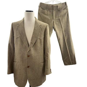 May include: A two-piece suit in a light brown tweed fabric. The jacket has a notched lapel, two front pockets, and two buttons. The pants are straight-legged with a high waist. The suit is displayed on a mannequin.