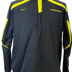Puede incluir: Una chaqueta tipo pullover Nike con cremallera de un cuarto en color negro, con detalles en amarillo brillante en el cuello, los hombros y las mangas. La palabra OREGON está impresa en amarillo en el pecho izquierdo y un logo Nike en amarillo.