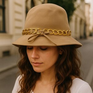 May include: A tan cloche hat with a gold chain detail around the crown and a small bow. The hat has a wide brim and a classic, elegant design. This hat is a stylish accessory for any outfit.