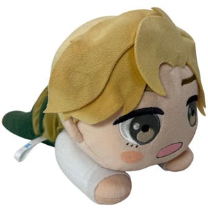 May include: A plush doll depicting a character with light blonde hair and a white shirt. The doll has large eyes and a surprised expression. The doll is lying down on a white surface.