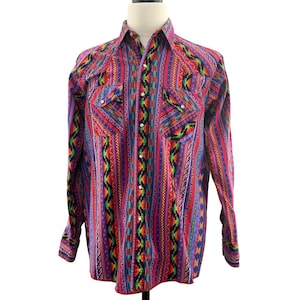 Puede incluir: Una camisa de manga larga con botones y un patrón vibrante y colorido. La camisa presenta una mezcla de rayas verticales y diseños geométricos en rojo, morado, azul, verde y amarillo. Tiene dos bolsillos en el pecho y botones blancos.