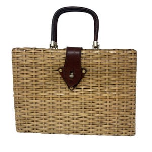 May include: A rectangular woven handbag with a dark brown leather handle and a matching leather flap closure. The bag is tan with silver-toned hardware and a small emblem on the flap. The handle is arched.