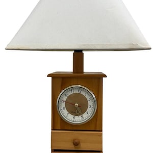 May include: A wooden table lamp with a white lampshade. The lamp features a clock with a silver bezel and a light brown wooden base with two small drawers. The clock face has black numerals.