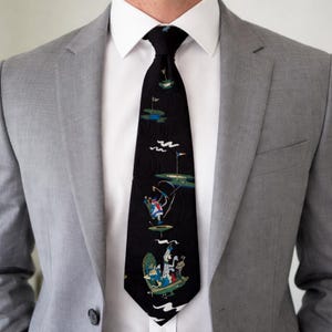May include: Black necktie with a golf-themed pattern. The tie features illustrations of golfers, golf holes, and flags in shades of blue, green, and white against a black background. The tie is worn with a white collared shirt and a gray blazer.
