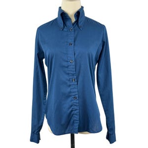 May include: A long-sleeved button-down shirt in a solid deep blue colour. The shirt features a pointed collar with button-down collar points, a central vertical button placket, and long sleeves with buttoned cuffs. The fabric has a smooth texture.