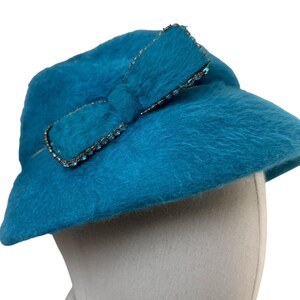 Puede incluir: Un sombrero azul turquesa con un lazo decorativo. El lazo está adornado con pequeños y brillantes pedrería. El sombrero tiene una textura suave y difusa y una forma de cloche clásica, perfecto para la moda de inspiración vintage.