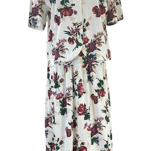 May include: A two-piece floral outfit comprising a short-sleeved button-up top and a midi skirt. The ensemble is white with a pattern of burgundy, purple, and green flowers. The top has a round neckline and the skirt has a gathered waist.