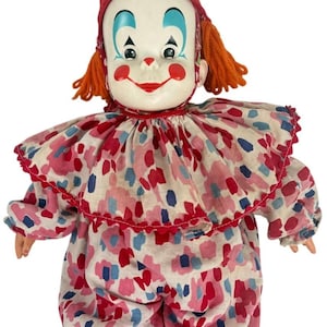 Pode incluir: Um boneco palhaço com rosto branco, sobrancelhas azuis pintadas, bochechas vermelhas e nariz vermelho. Usa um chapéu de tecido vermelho com um pompom branco, cabelo de lã laranja, e um colarinho com babados e macacão com padrão abstrato rosa, vermelho e azul.