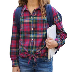 Puede incluir: Una camisa de franela a cuadros roja, verde, amarilla y azul está atada a la cintura. La persona lleva vaqueros azules y una mochila azul marino. Un cuaderno de espiral blanco se sostiene en la mano de la persona.