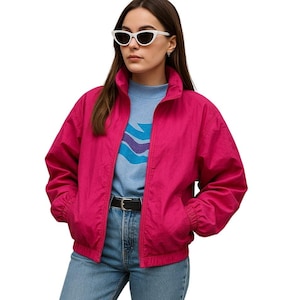 May include: A bright pink, zip-up jacket with a high collar. The jacket is paired with a light blue sweater featuring a wave design, blue jeans, and white sunglasses. A black belt cinches the waist.