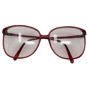 May include: Oversized burgundy eyeglasses with a rounded rectangular frame. The clear lenses are set in a thin frame. The temples extend to the ear pieces. The glasses have a vintage aesthetic.