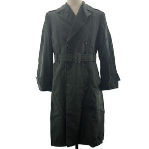 May include: A dark charcoal gray double-breasted trench coat featuring a wide notched collar, shoulder epaulets, and a matching fabric belt with a rectangular buckle. The coat has long sleeves with buttoned cuffs and six front buttons.