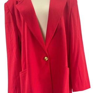Op de afbeelding: Een felrode blazer met een enkele goudkleurige knoopsluiting in het midden, brede revers en twee voorzakken. Het kledingstuk heeft lange mouwen en een gestructureerd silhouet, getoond op een etalagepop tegen een witte achtergrond.