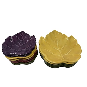 Può includere: Una collezione di piatti in ceramica a forma di foglia in due dimensioni distinte. La pila più piccola presenta piatti viola, gialli, rossi e verdi. La pila più grande è composta da piatti gialli con dettagli venati visibili sulla superficie.