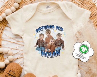 May include: A natural colored baby onesie with a blue and white graphic that says "Birthday Boy Nolan" with a photo of four young boys. The onesie has a green organic cotton certification tag.