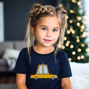 May include: A young girl with blonde hair and blue eyes wearing a navy blue t-shirt. The shirt features a yellow cartoon car design. The background includes a blurred Christmas tree with warm lights.