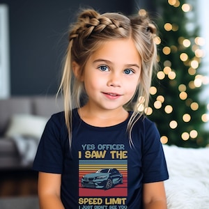 May include: A young girl wearing a navy blue t-shirt with the text "YES OFFICER I SAW THE SPEED LIMIT I JUST DIDN'T SEE YOU." The shirt features a car graphic with a retro color scheme.
