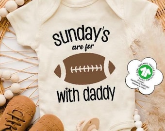 May include: An off-white short-sleeve baby bodysuit featuring the black text "sunday's are for with daddy" and a brown football graphic. A white flower-shaped GOTS organic textile certification label is attached to the side of the garment.