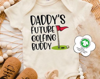 May include: An off-white short-sleeve baby bodysuit featuring the black text DADDY'S FUTURE GOLFING BUDDY, a red golf flag on a pole, and a small white golf ball on a green patch. A white flower-shaped GOTS organic textile certification tag is attached.