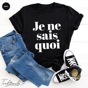 May include: Black t-shirt with white text that reads "Je ne sais quoi". The shirt is laid flat on a white wooden surface with a pair of blue jeans and black sneakers.