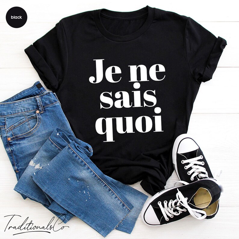 May include: Black t-shirt with white text that reads "Je ne sais quoi". The shirt is laid flat on a white wooden surface with a pair of blue jeans and black sneakers.