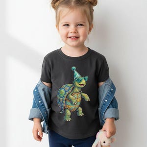 May include: A charcoal gray t-shirt featuring a colorful cartoon turtle wearing sunglasses and a party hat. The turtle has a patterned shell and is smiling. The shirt is worn by a young child.