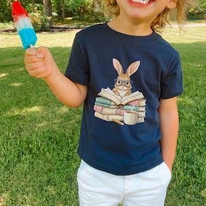 May include: Navy blue t-shirt featuring a cartoon rabbit wearing glasses and reading a book. The illustration includes a stack of books, a cup of coffee, and a pencil. The child is holding a red, white, and blue popsicle.