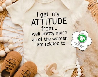 May include: An off-white short-sleeve baby bodysuit featuring the black text: I get my ATTITUDE from... well pretty much all of the women I am related to. The garment includes a GOTS organic certification tag and is displayed with brown shoes.