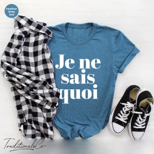 May include: A blue t-shirt with the French phrase "Je ne sais quoi" printed in white on the front. The shirt is laid flat on a white wooden surface with a black and white plaid shirt and black sneakers.