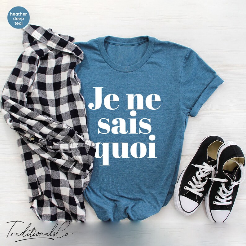 May include: A blue t-shirt with the French phrase "Je ne sais quoi" printed in white on the front. The shirt is laid flat on a white wooden surface with a black and white plaid shirt and black sneakers.