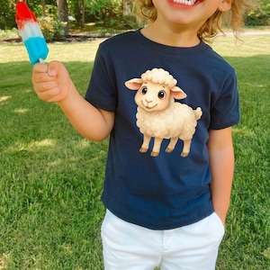 May include: Navy blue t-shirt featuring a cartoon sheep illustration. The sheep is beige with a fluffy head and smiling face. The child is also holding a red, white, and blue popsicle. The child is wearing white shorts.