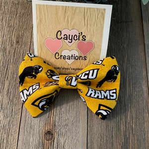 May include: Yellow bow tie with a black and white print of a ram's head and the words "Rams" and "WCU". The bow tie is on a wooden surface. The text "Cayci's Creations" and a website address are visible on a card behind the bow tie.
