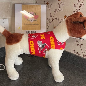 May include: A red dog diaper with a repeating pattern of the Kansas City Chiefs logo and the word "CHIEFS" in yellow. The diaper is on a stuffed dog with brown and white fur. A sign in the background reads "Luci's Creations".