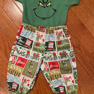 May include: A green short-sleeved bodysuit featuring a black graphic of The Grinch's face, paired with pants covered in a multi-colored grid pattern of Dr. Seuss themed text and illustrations including Who Hash cans and holiday motifs.
