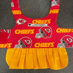 May include: A red and gold pet accessory featuring the Kansas City Chiefs logo and helmet design. The accessory has a yellow pleated skirt and the word "CHIEFS" in gold. The accessory has a velcro closure.