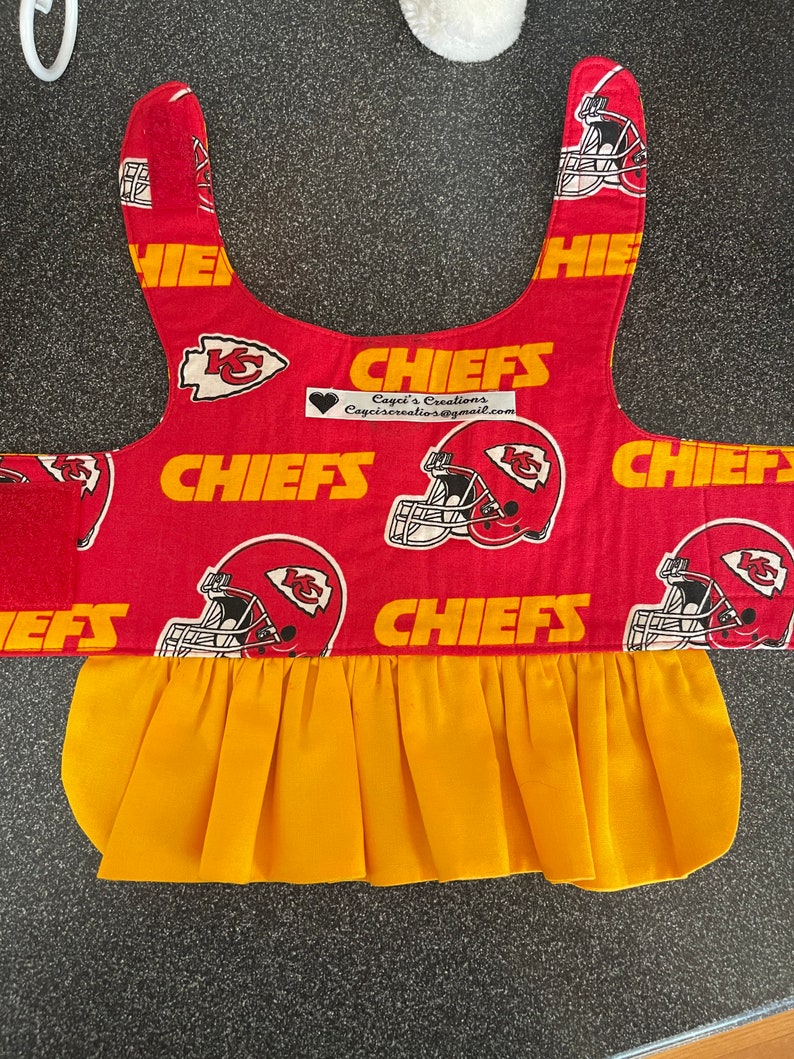 May include: A red and gold pet accessory featuring the Kansas City Chiefs logo and helmet design. The accessory has a yellow pleated skirt and the word "CHIEFS" in gold. The accessory has a velcro closure.