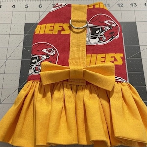 May include: A red and yellow dog dress featuring the Kansas City Chiefs logo and text. The dress has a yellow bow and a pleated skirt. The top portion of the dress is red with the team's logo and the word "CHIEFS" in yellow.
