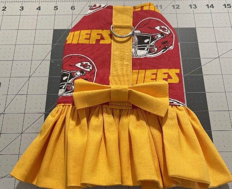May include: A red and yellow dog dress featuring the Kansas City Chiefs logo and text. The dress has a yellow bow and a pleated skirt. The top portion of the dress is red with the team's logo and the word "CHIEFS" in yellow.