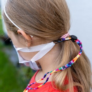 Könnte beinhalten: Eine junge Person trägt eine weiße Gesichtsmaske mit einem bunten Lanyard um den Hals. Das Lanyard besteht aus gewebtem Stoff in verschiedenen Farben, darunter Rot, Orange, Gelb, Grün, Blau, Lila und Pink.