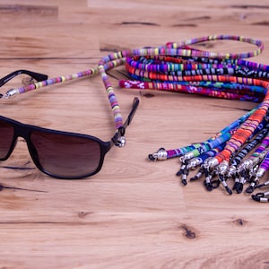 May include: A set of colorful woven eyeglass straps with silver metal clasps. The straps are made of a variety of colors and patterns, including red, blue, green, yellow, and black. The straps are shown on a wooden surface.